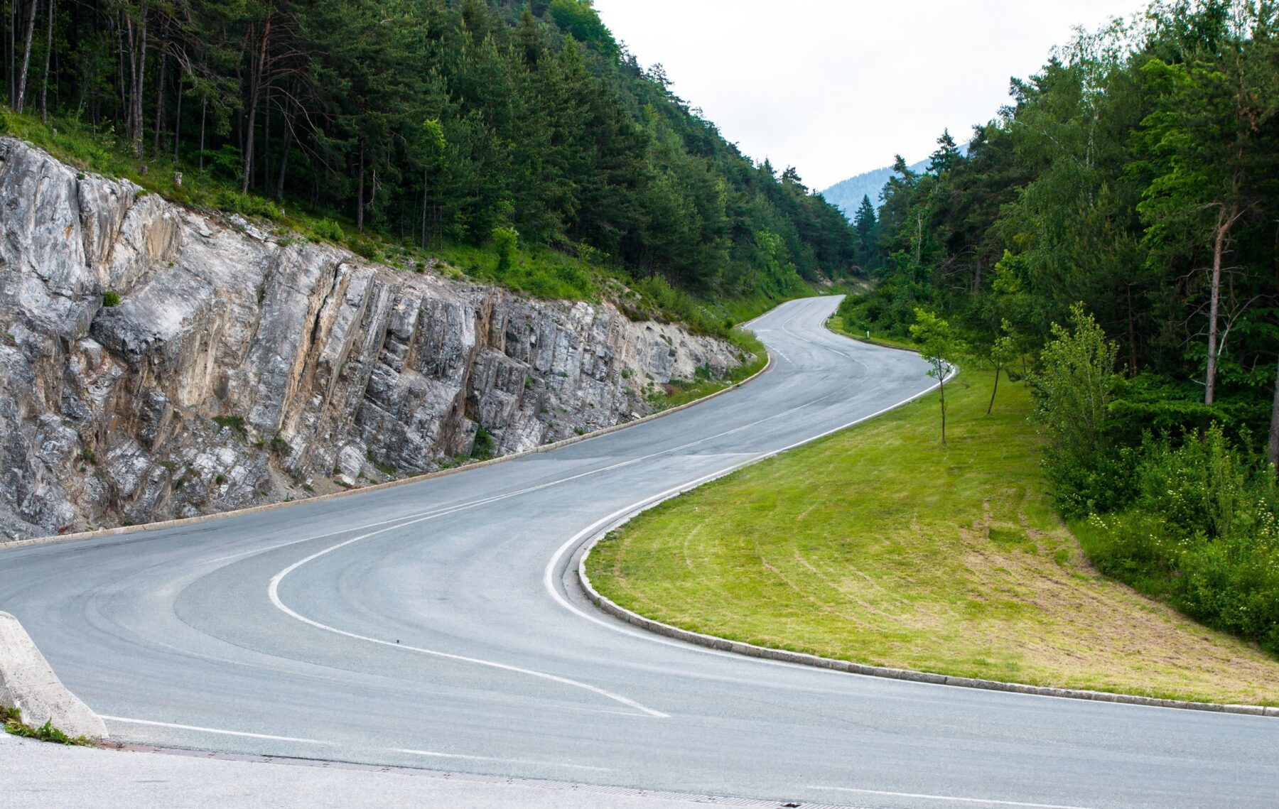 Winding asphalt mountain road with sharp curves, surrounded by rocky cliffs and dense forest.